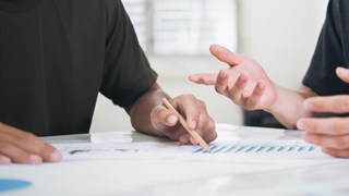 Detalj på två personer som sitter vid ett bord med papper och pratar med varandra. För att illustrera samarbete, möte eller grupparbete.