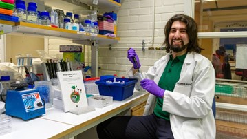 En man som sitter i ett laboratorium klädd i labbrock och handskar. Han har långt mörkt hår och skägg.