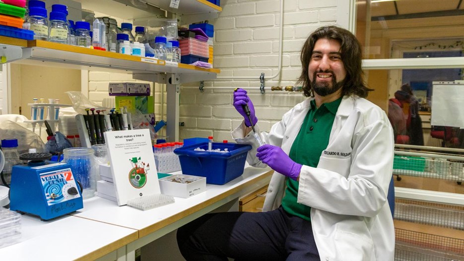 En man som sitter i ett laboratorium klädd i labbrock och handskar. Han har långt mörkt hår och skägg. 