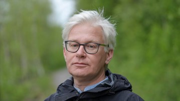 En man med glasögon som står på en grusväg.