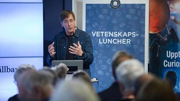 Föreläsare pratar inför publik, rollup med ordet vetenskapsluncher syns i bakgrunden.