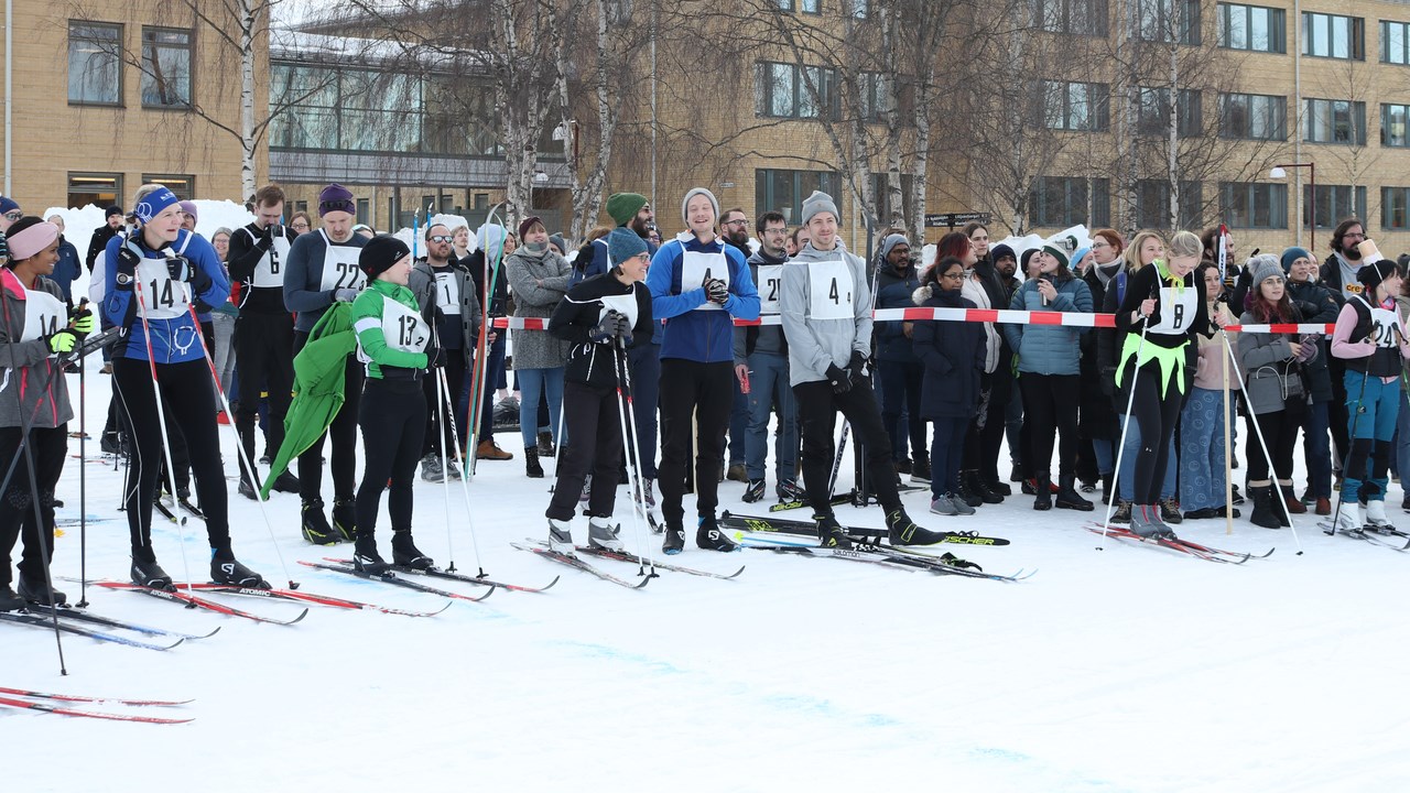 Several people on skis with numbers on their breast are waiting in front of  a white and red plastic band for their turn in the skiing relay. Behind of the plastic band, people cheering for the teams are standing.