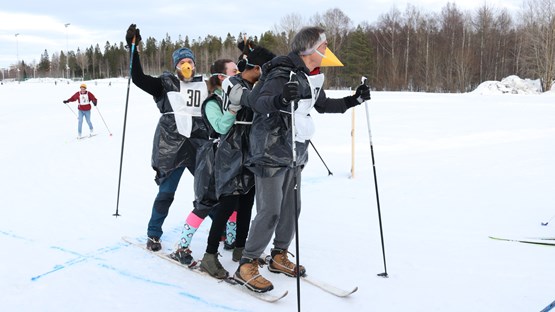 Four people skiing on one pair of long skis are crossing the finish line. They are dressed up with a yellow peak in front of their face. The first and the last person are holding sticks. 