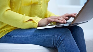 Detaljbild på kvinna i soffa som sitter med uppslagen laptop i en soffa och skriver.