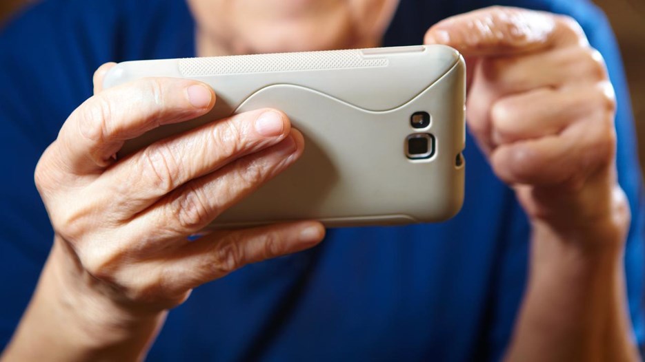 En äldre dam sitter med händerna  och navigerar en mobiltelefon. Det är endast händerna och mobiltelefonen som syns.