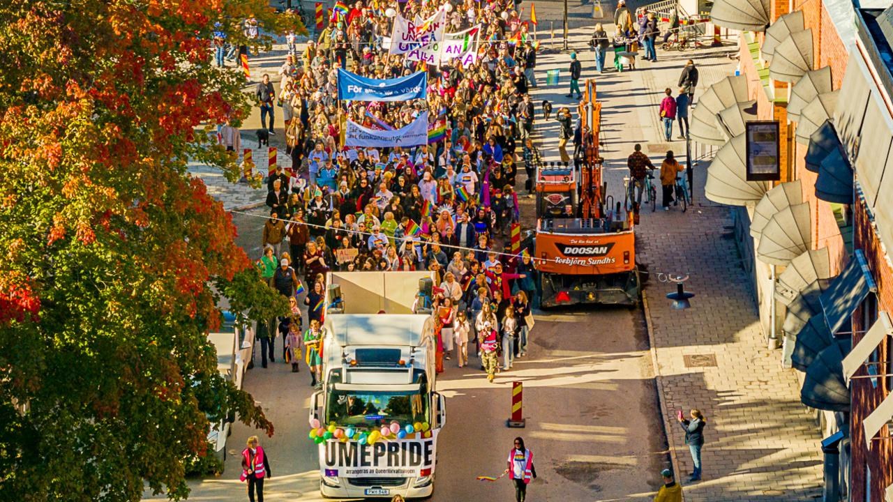Drönarbild av en parad av människor som går i Pride-tåget i Umeå 2024, med regnbågsflaggor och banderoller.