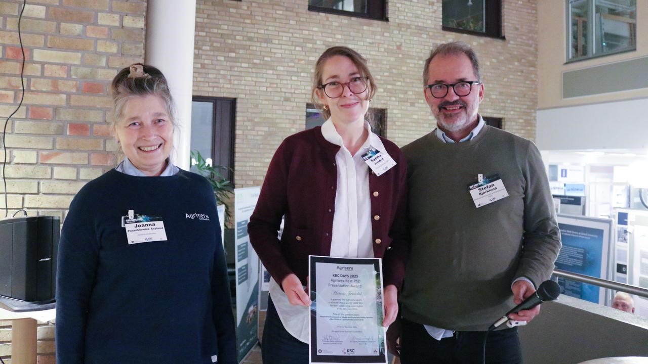 Three people are standing in the conference venue, the woman in the middle is holding an award certificate