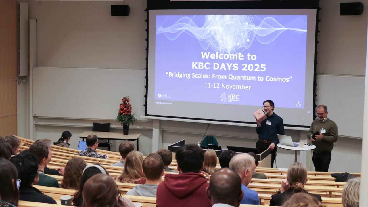Participants of the scientific conference are listening to the presenter, chairperson and the presenter are standing by the digital screen