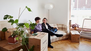 En man och en kvinna sitter i en soffa i ett vardagsrum, omgivna av flyttkartonger. 