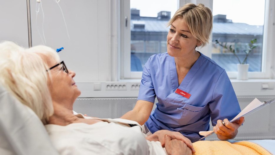 A nurse is sitting next to a bed where an elderly woman is lying