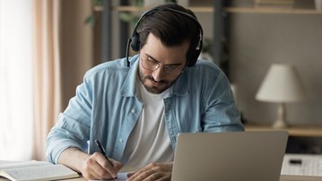 En man sitter vid ett bord med laptop och headset, böcker och antecknar i ett block.
