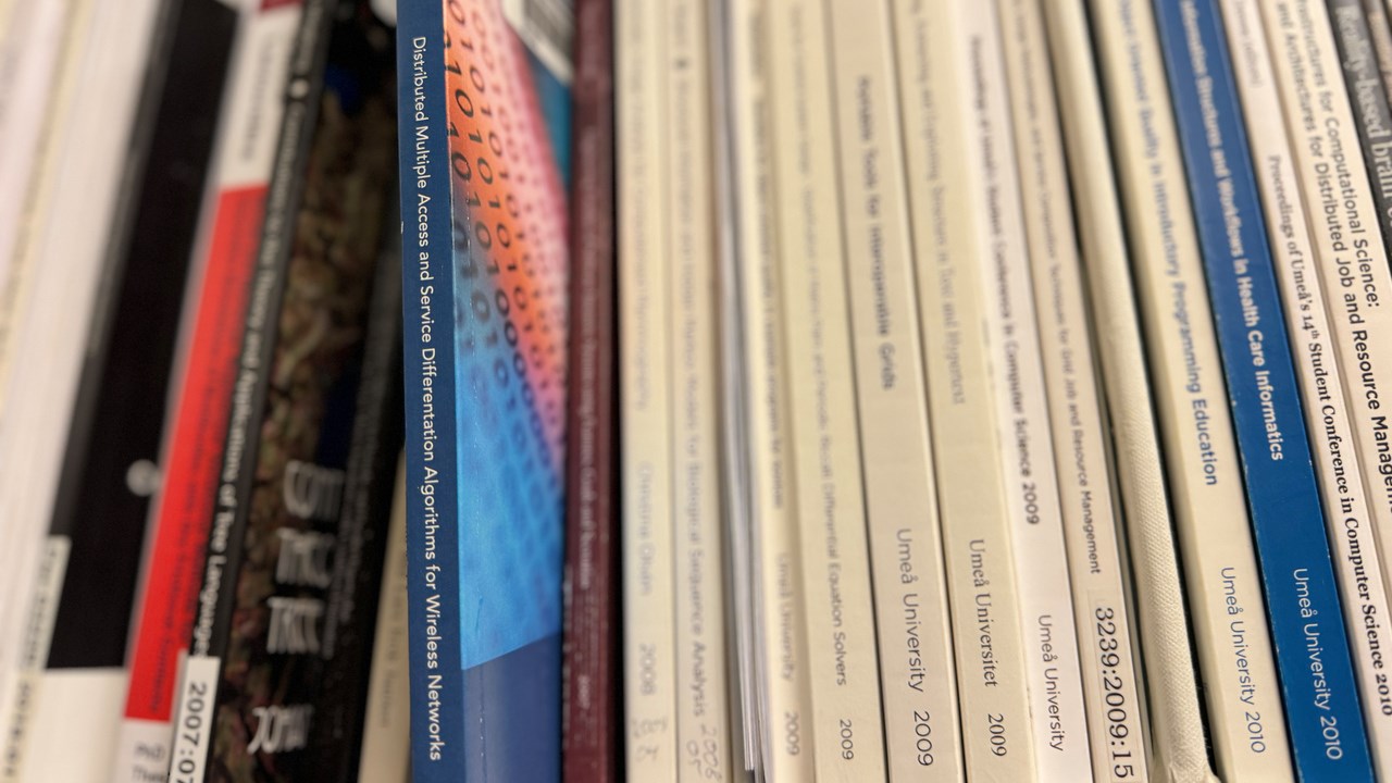 Thesis among other books in the university library. The book is blue in colour with red elements on the front cover.