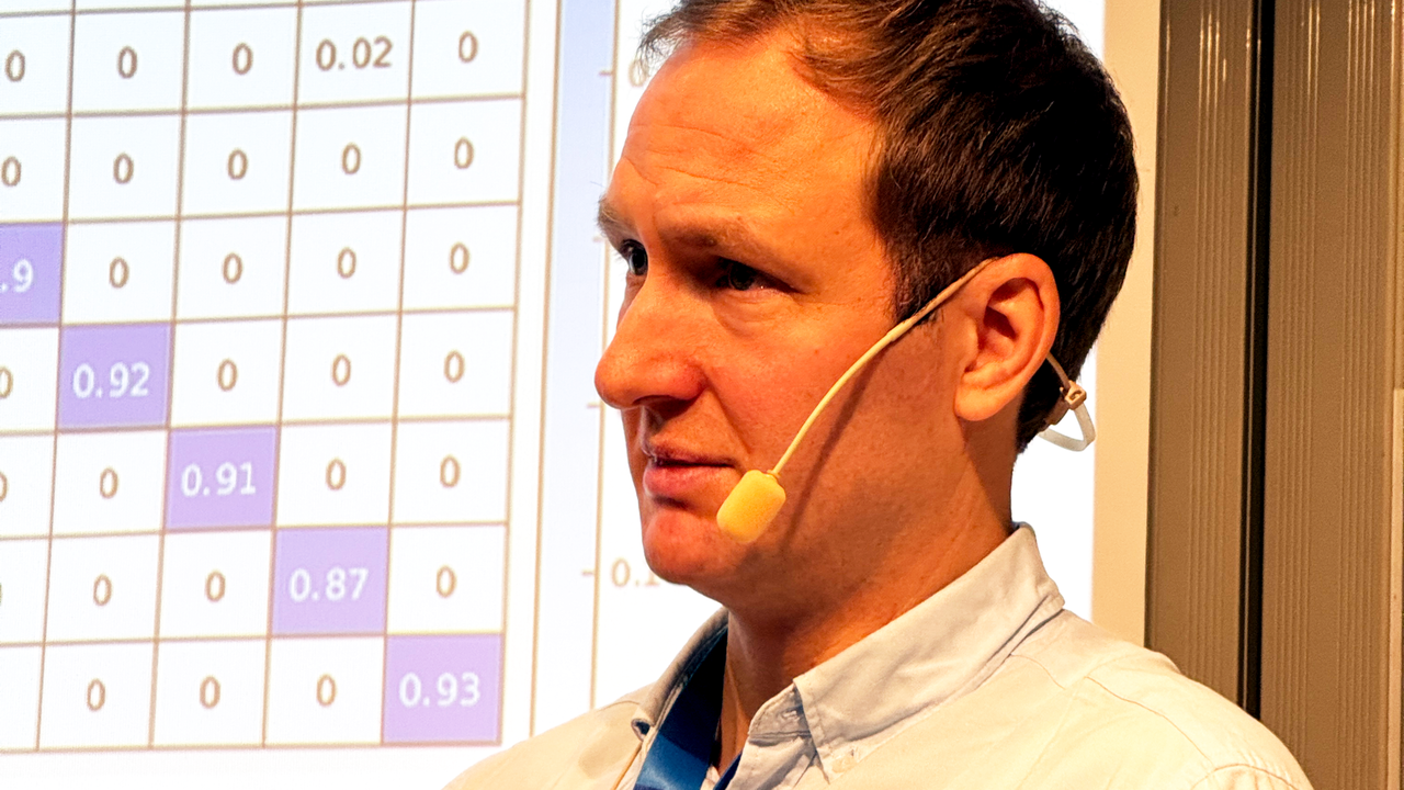 Man in front of a projection screen showing a mathematical matrix. He is wearing a light blue shirt and has a microphone clip around his ear.