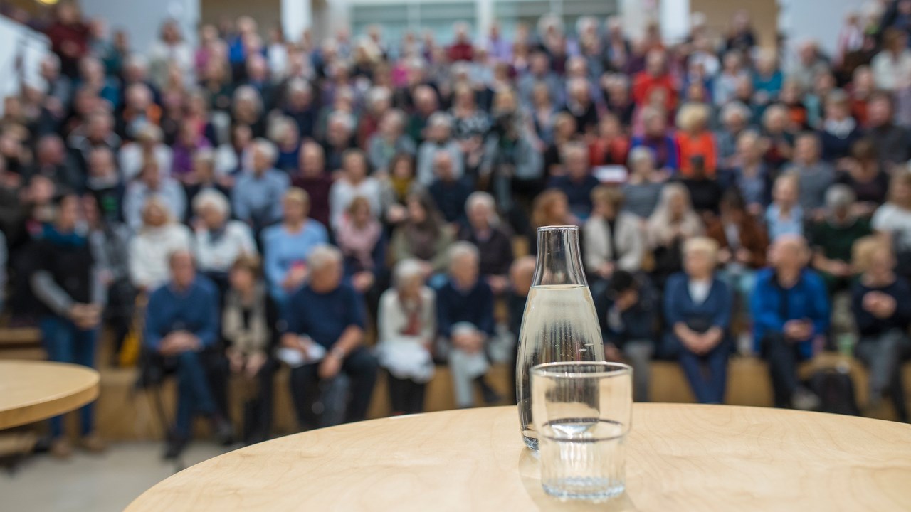 Bild på vattenkaraff med glas på ett bord framför en publik. Bilden ska illustrera att hålla föredrag.