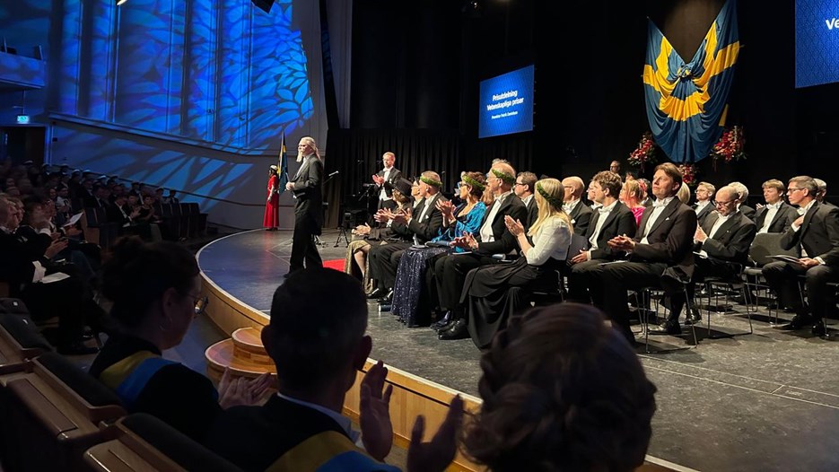 En upplyst scen med folk som sitter på stolar. En man står längst fram och tar emot applåder.