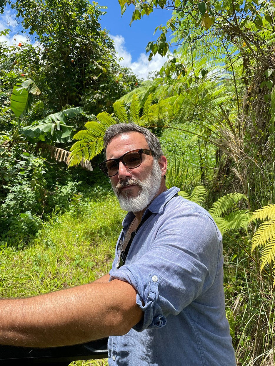 Luis Berrios-Negrón stands in the foreground with a blue shirt and sunglasses.  He is surrounded by a green rain forest and blue skies.