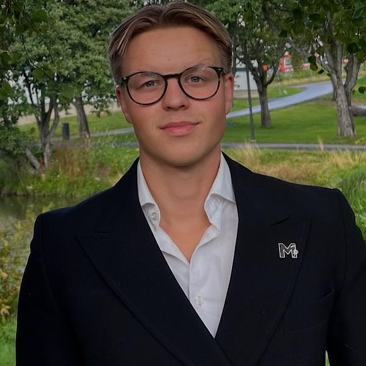 En man i kostym st&aring;r bredvid ett tr&auml;d.