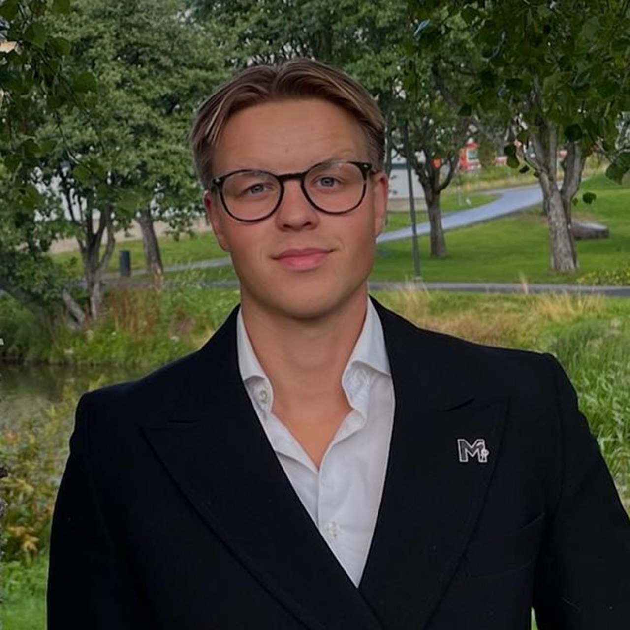 En man i kostym st&aring;r bredvid ett tr&auml;d.