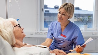 Sköterska sitter bredvid en säng där en äldre kvinna ligger