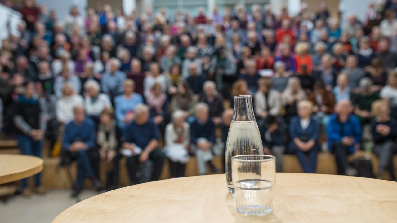 Bild på vattenkaraff med glas på ett bord framför en publik. Bilden ska illustrera att hålla föredrag.