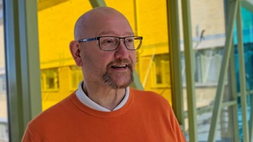 Man utan hår, med svarta glasögon, en orangetröja med vid skjorta under, som står framför ett fönster.