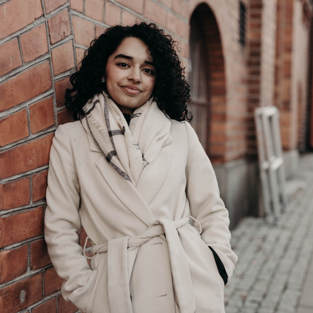 Ung kvinna med mörkt lockigt hår och beige kappa står inne i Umeå centrum framför en vägg och ler.