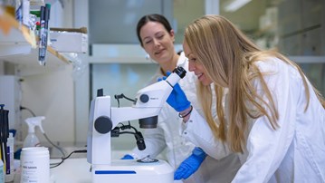 Two women in lab coats looking through a microscope.