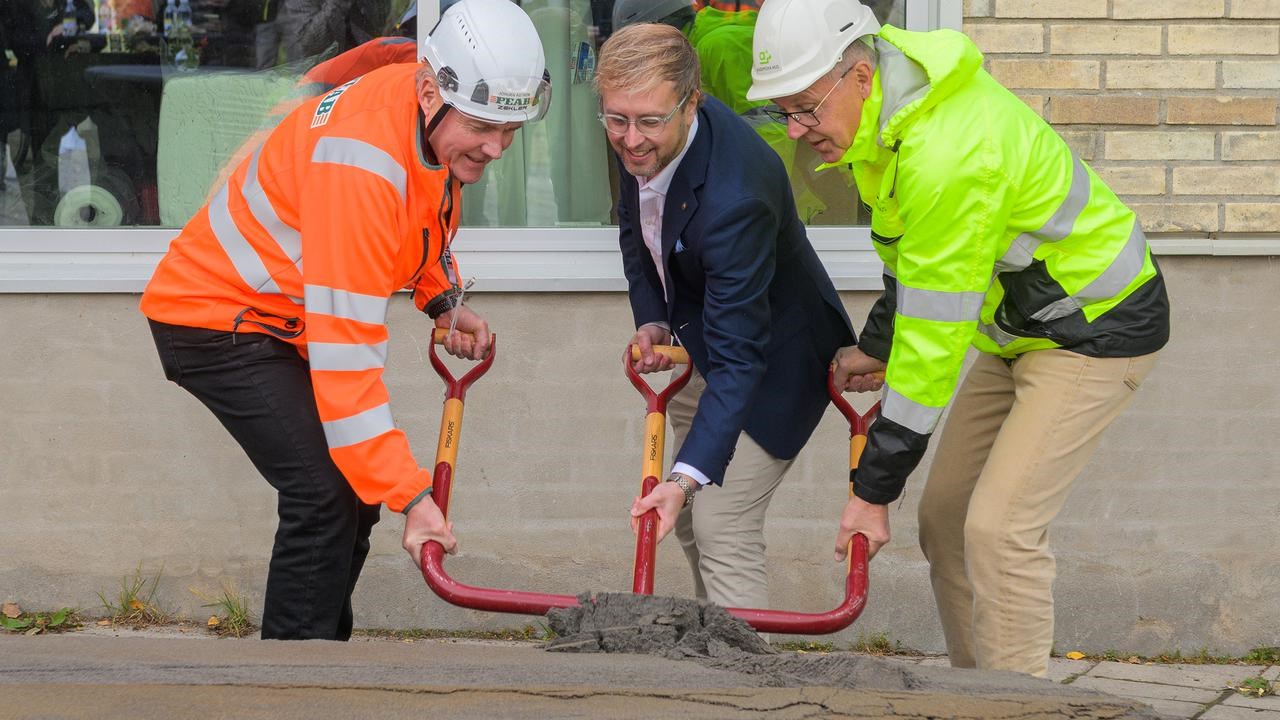 Tre m&auml;n som st&aring;r bredvid varandra och tar ett gemensamt spadtag med specialbyggd spade med tre skaft framf&ouml;r en byggnad.
