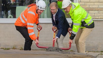 Tre män som står bredvid varandra och tar ett gemensamt spadtag med specialbyggd spade med tre skaft framför en byggnad.