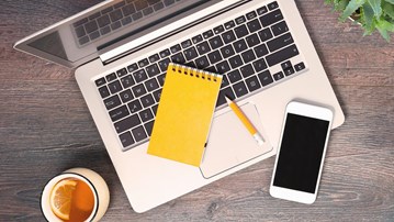 Dator, anteckningsblock, mobil och penna fotat uppifrån på ett bord.