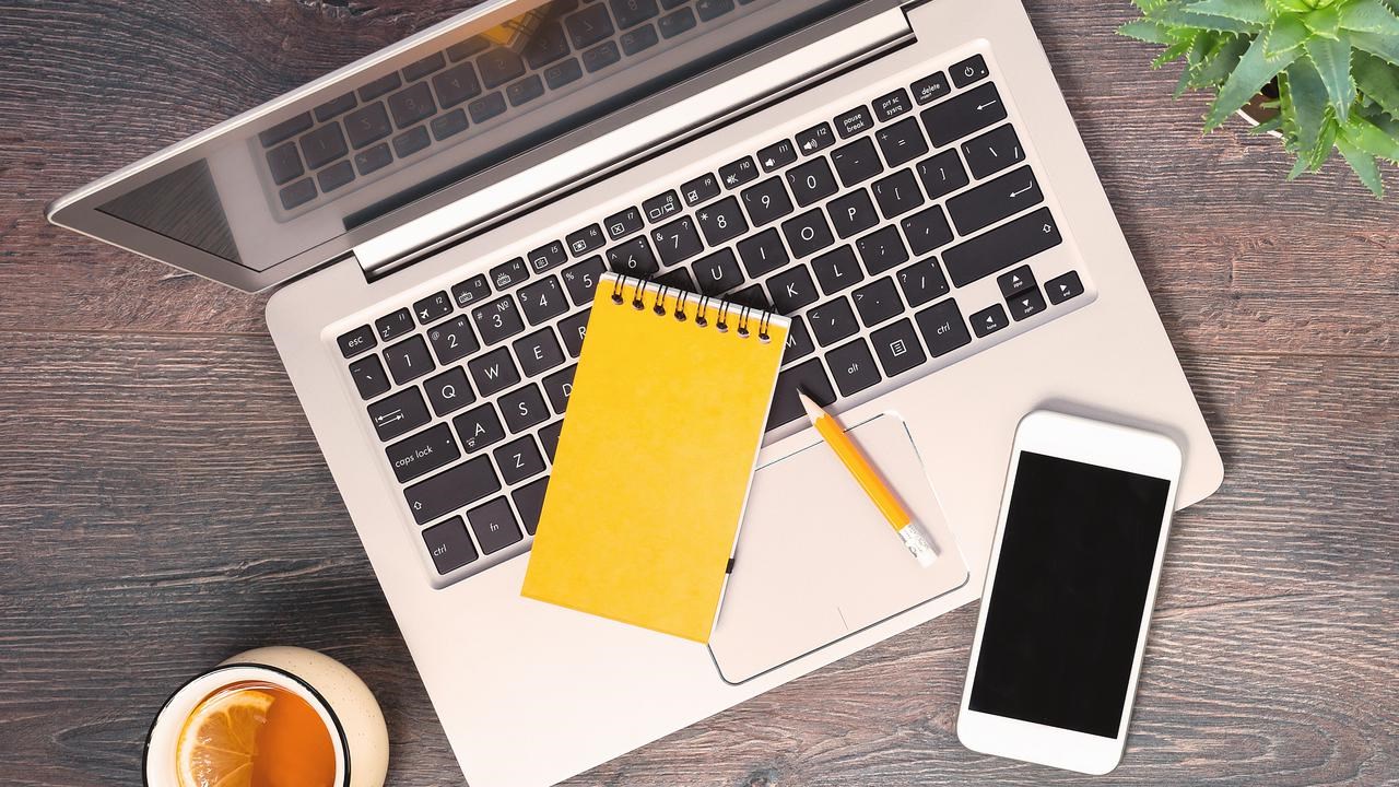 Dator, anteckningsblock, mobil och penna fotat uppifrån på ett bord.