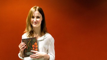 En kvinna med långt brunt hår och vit skjorta håller en bok med brunt-rött omslag i handen. Hon står framför en röd vägg.