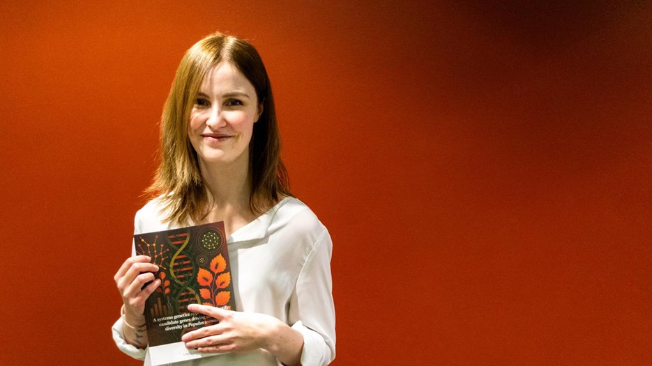 En kvinna med långt brunt hår och vit skjorta håller en bok med brunt-rött omslag i handen. Hon står framför en röd vägg.