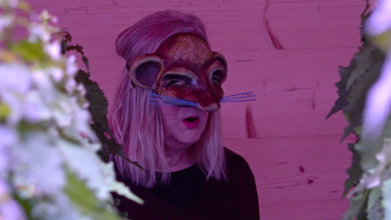 A blonde woman with a black t-shirt a face mask looking like a mouse, sits on a stage with pink light, and some hand made trees in the foreground.
