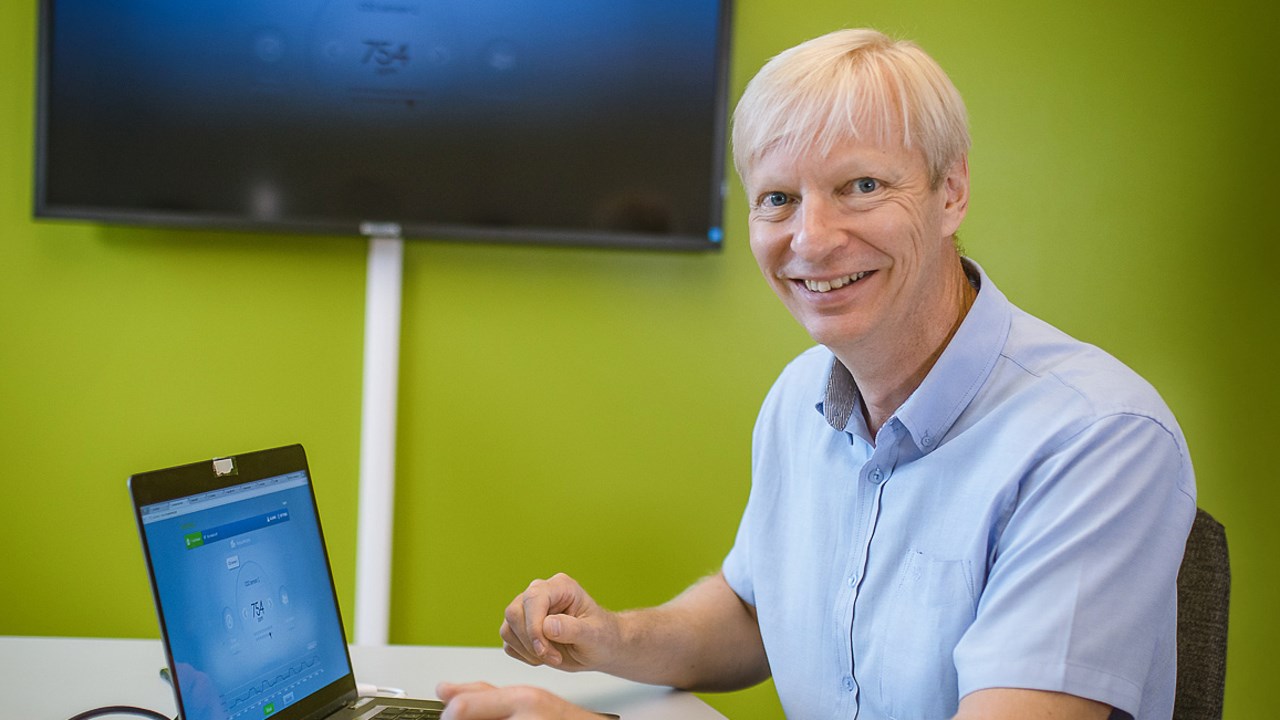 Professor Kary Främling sitter vid ett skrivbord med dator och en skämr på väggen. Han är blond och ler mot kameran i en ljusblå kortärmad skjorta.