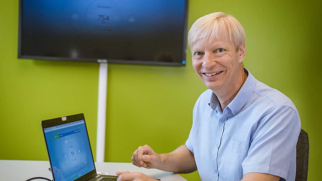 Professor Kary Främling with a computer on the desk and a screen on the wall, is smiling to the camera.