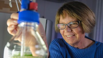 En kvinna i närbild med handen på en laboratorieflaska i förgrunden.