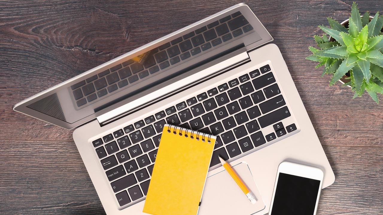 Dator, anteckningsblock, mobil och penna fotat uppifrån på ett bord.