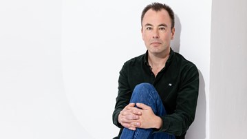 Marcus Gustafsson sitter med knät uppdraget och armarna runt benet, han tittar rakt in i kameran. Han har jeans och en svart skjorta på sig. 