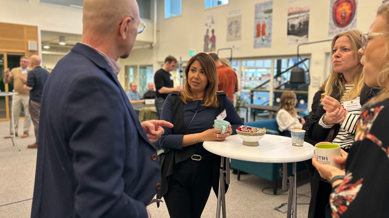 Fyra personer står samlade kring ett bord och pratar med människor minglande i bakgrunden.