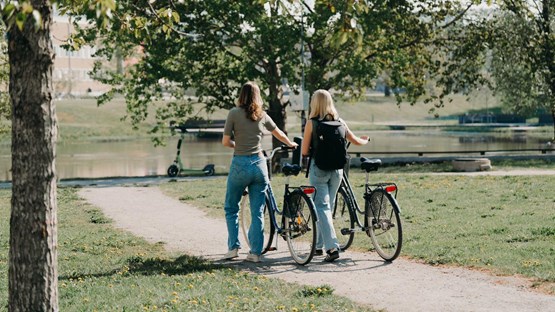 Två kvinnor går bredvid varandra på en grusväg och leder varsin cykel.