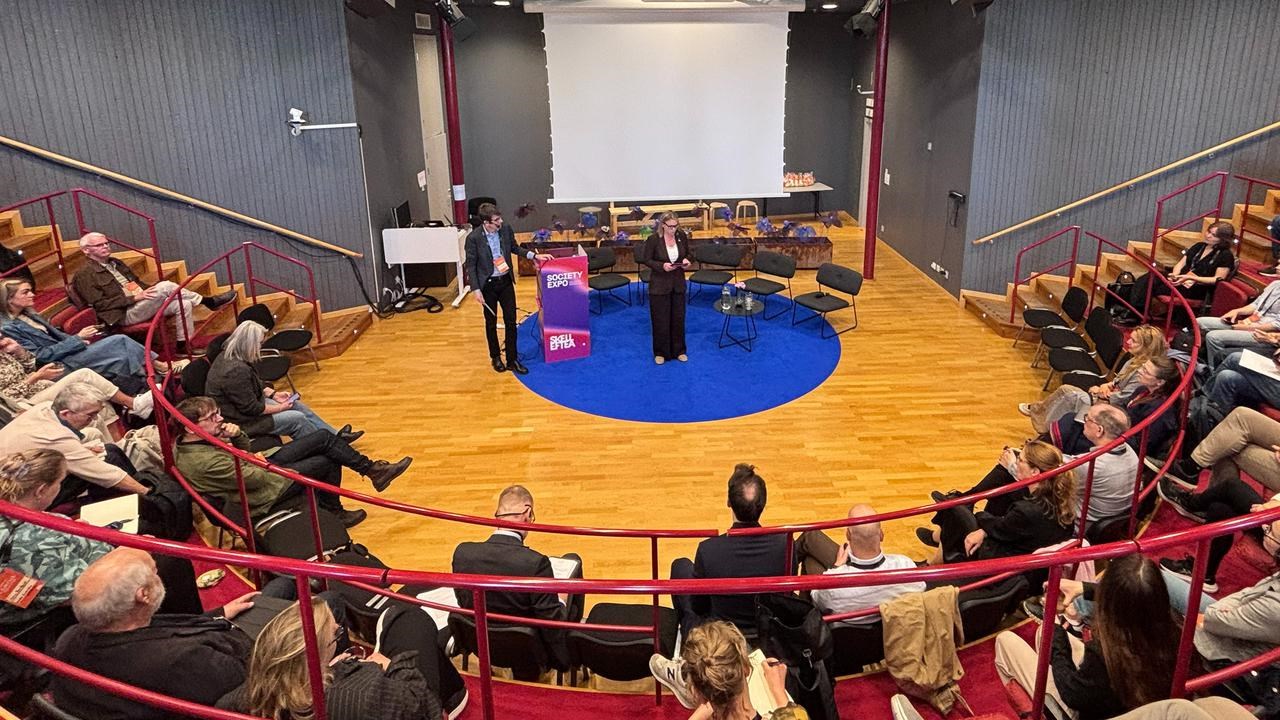 En stor publik tittar p&aring; ett par talare som st&aring;r p&aring; scen.