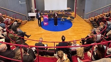En stor publik tittar på ett par talare som står på scen.