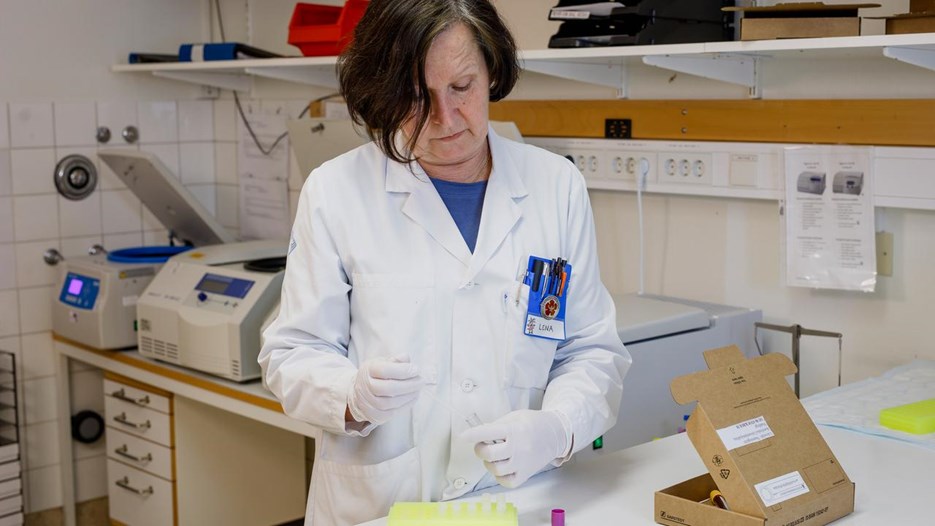Lena Bylund står vid en labbänk och pipetterar ur ett provrör. På bänken står ett provrörsställ och en öppen kartong som anlänt till kliniken. I bakgrunden syns centrifuger på en annan labbänk.