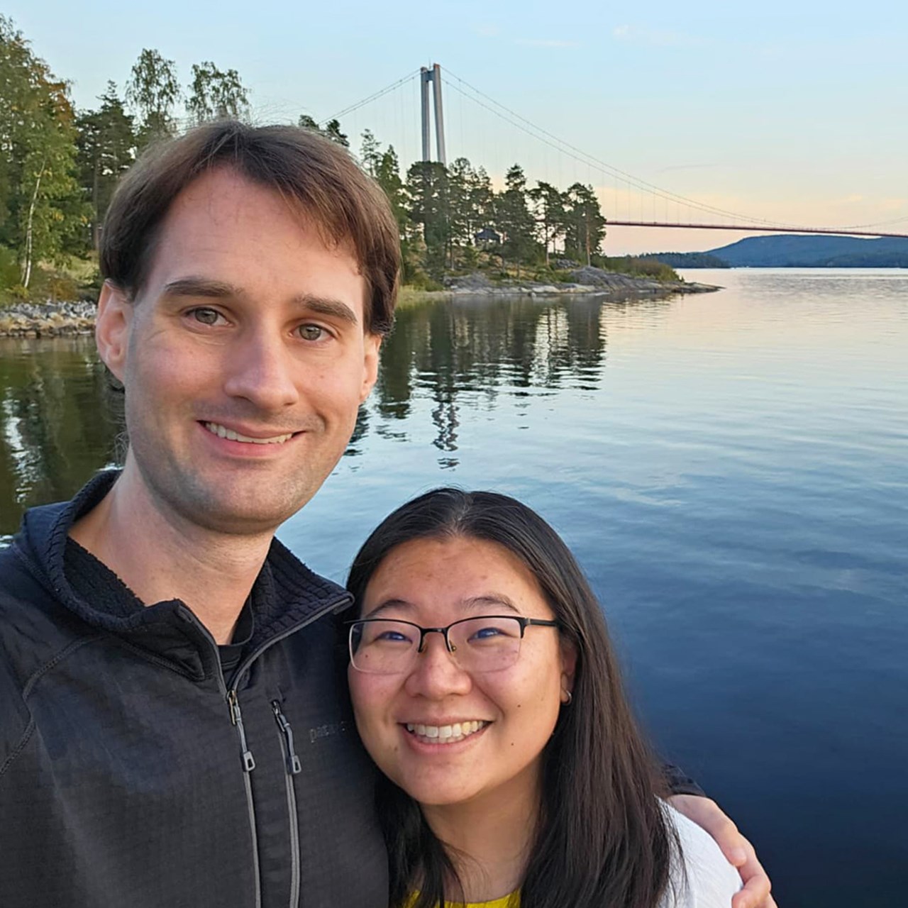 Par som tar selfie framför Höga kusten-bron i solnedgång.