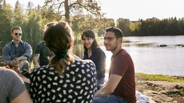 A group of people sat around a campfire by a lake in the evening sun.