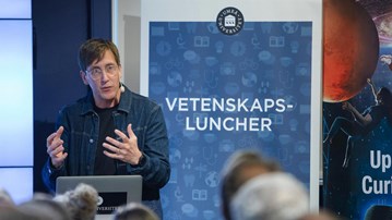 Föreläsare pratar inför publik, rollup med ordet vetenskapsluncher syns i bakgrunden.