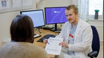 I ett undersökningsrum sitter en person med ryggen mot kameran och tittar på ett formulär som Ivar Winroth håller upp. I bakgrunden syns ett skrivbord med datorskärmar.