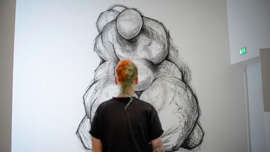 A person stands with the back facing the camera and a big drawing on the wall in front.