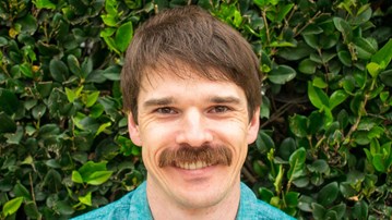 A man, wearing a blue shirt, with a rich moustache and smile on hisface is standing in front of a thick, bright green hedge.